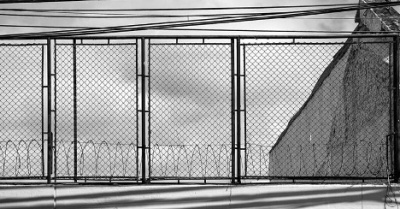 Fotografía que muestra un portón de hierro y malla formando líneas con cables en la parte superior y su sombra además de alambre de púas en la parte inferior